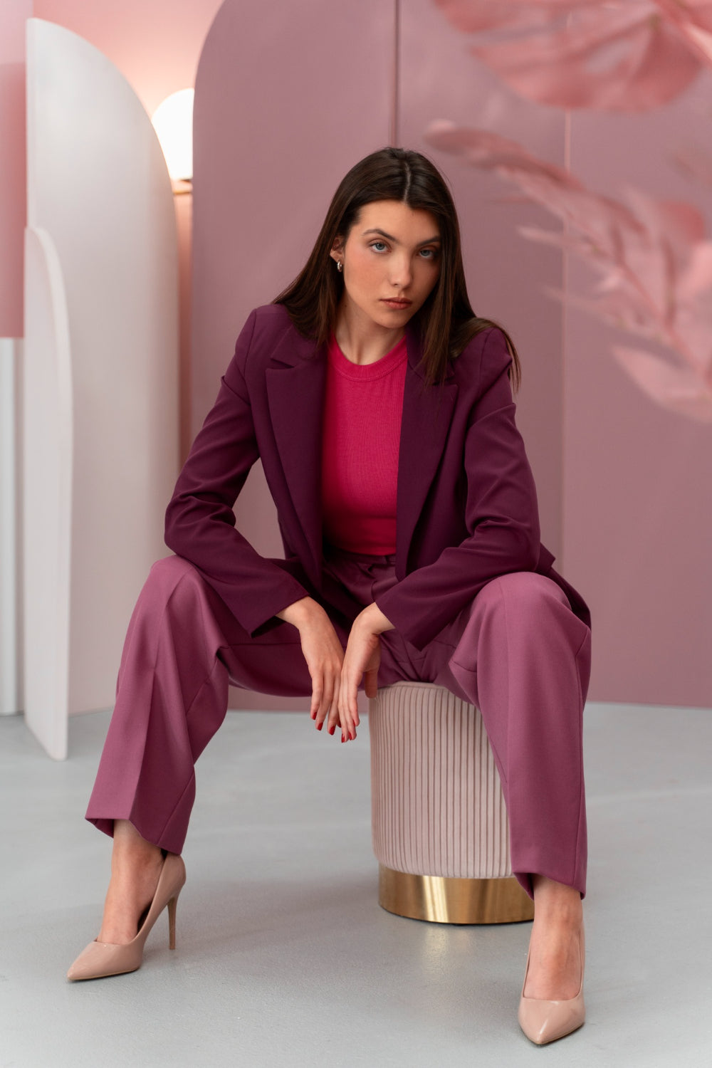 Woman in a purple suit sitting on a stool against a pink background
